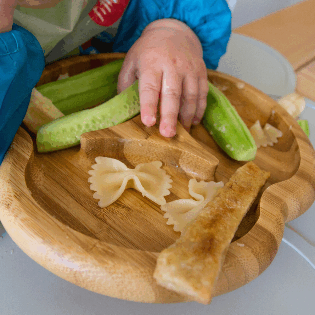 Baby moving their hand across their plate of finger foods to pick up cooked, soft pasta with their palmar grasp.