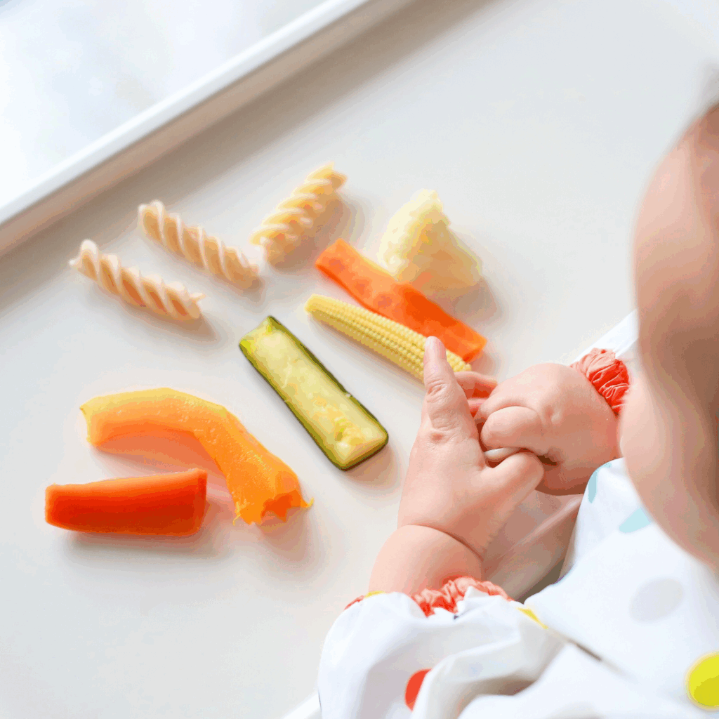 A baby sits at their high chair with a number of finger food options on the tray, including pasta, cooked carrot, zucchini, and peppers.