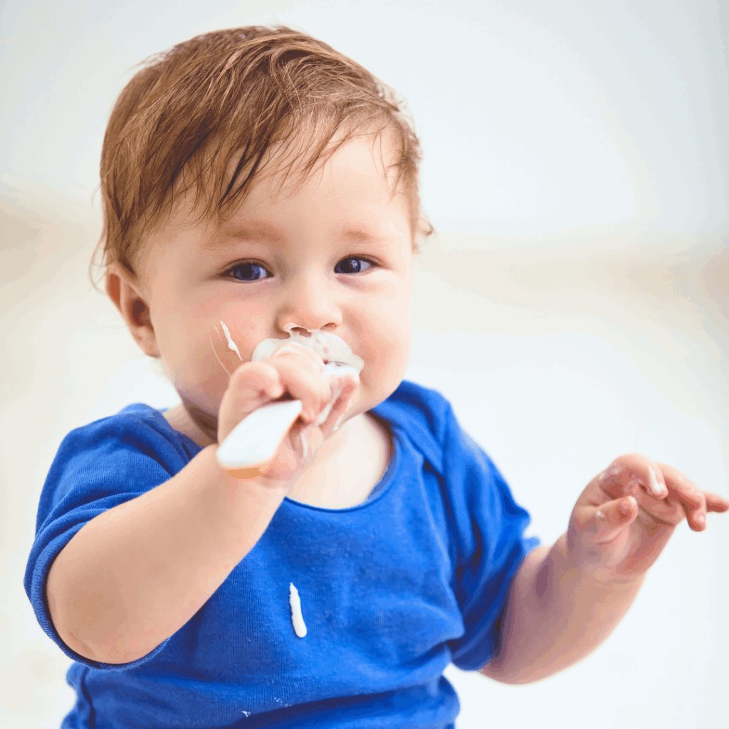 A baby self-feeds with a spoon, eating yogurt at mealtime.