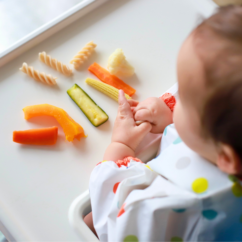 Top view of a baby sitting in a high chair exploring a variety of foods and textures including soft cooked carrots, squash, zucchini, cauliflower, corn, and plain pasta.