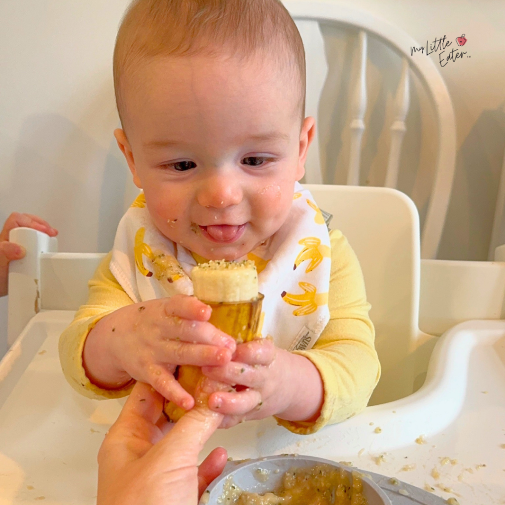 6 month old baby sitting in a high chair reaching out to grab a banana pop with hemp seeds from his mom with 2 hands, while smirking, tongue sticking out and eyes looking down at the banana.