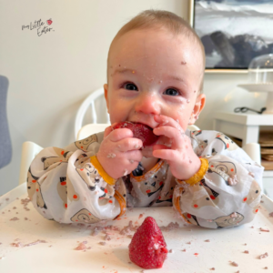 6 month old baby sitting in high chair eating a whole ripe strawberry with two hands while smirking.