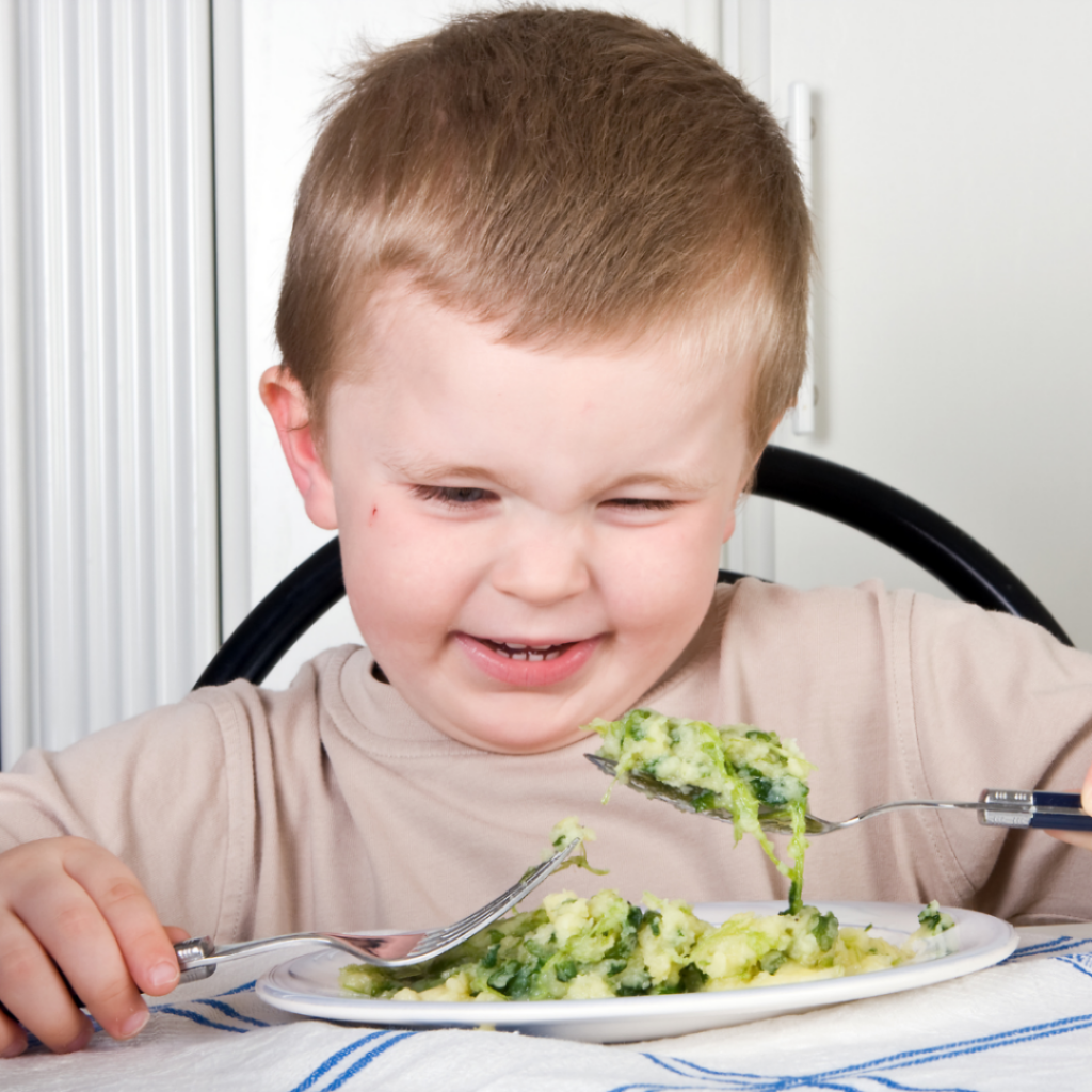Toddler sitting at the table making a funny face at a big spoonful of green veggies.