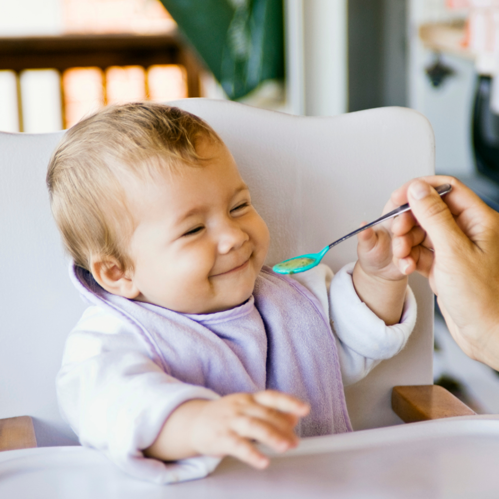 Baby in high chair smiling as she reaches out for a spoon full of pureed food.
