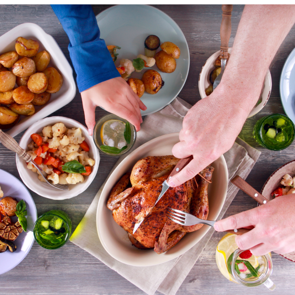 Top view of a family style meal with food layed out on the table, man cutting into a roasted chicken with side dishes all around