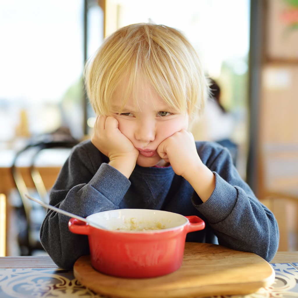 Child sitting at the table with a red bowl in front of him, showing disinterest in eating