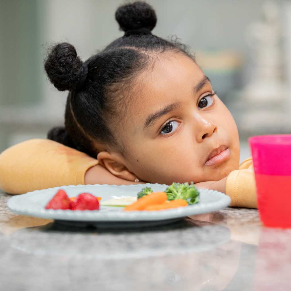 Toddler with head lying on her hands looking at a plate of food with veggies, showing disinterest in eating.