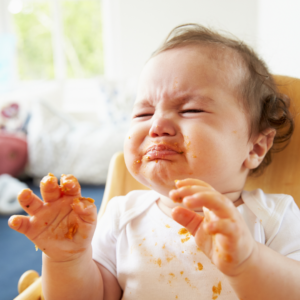 Baby with food on hands and face, scrunching her face and lips closed tightly.