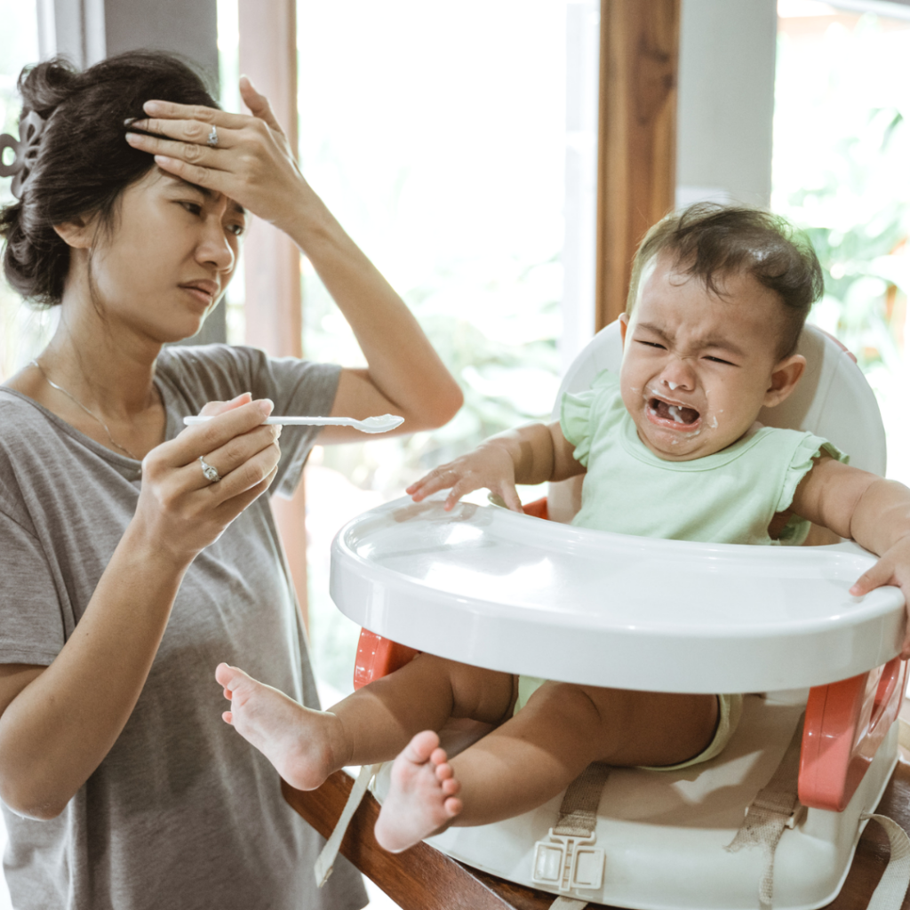 Baby crying in high chair with yogurt in his mouth; mom holding a spoon with hand on her head showing stress.