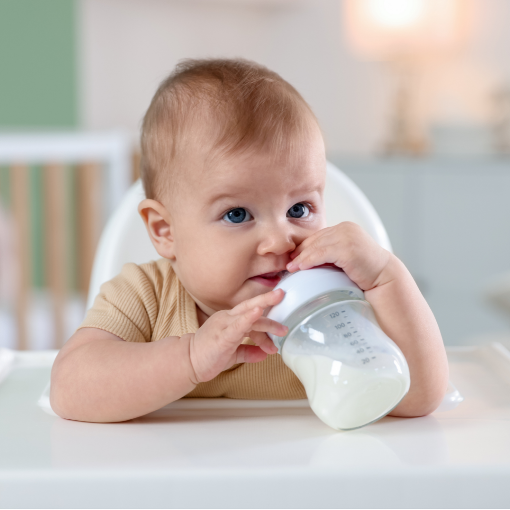Baby sitting in high chair holding a bottle of milk up to their mouth.