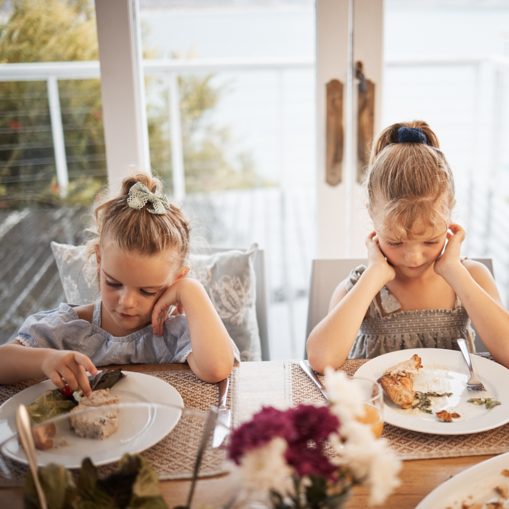Two toddlers sitting at the dinner table with plates of food, showing disinterest in eating.