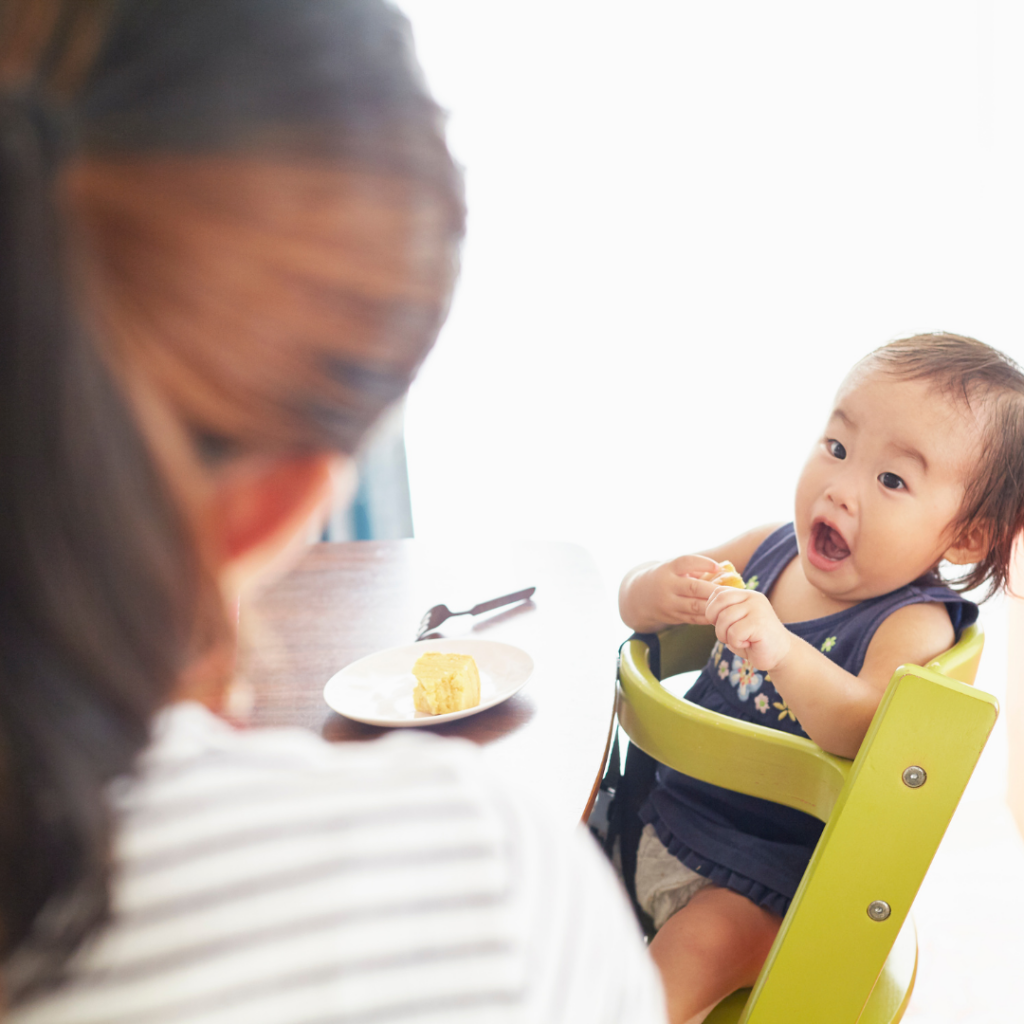 Baby sitting in high chair looking at mom while holding a piece of food with her mouth open.