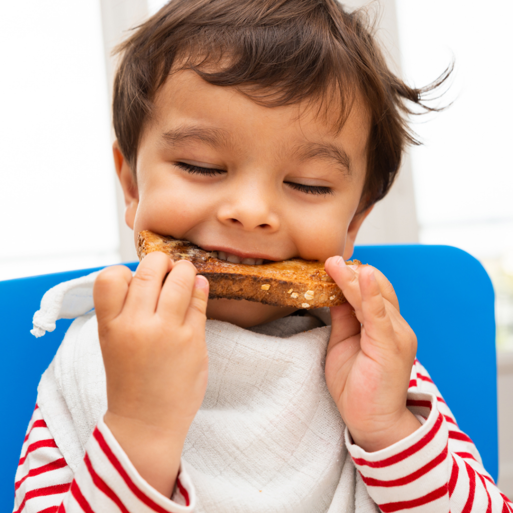Toddler taking a bite of whole grain toast while self-feeding in a high chair.